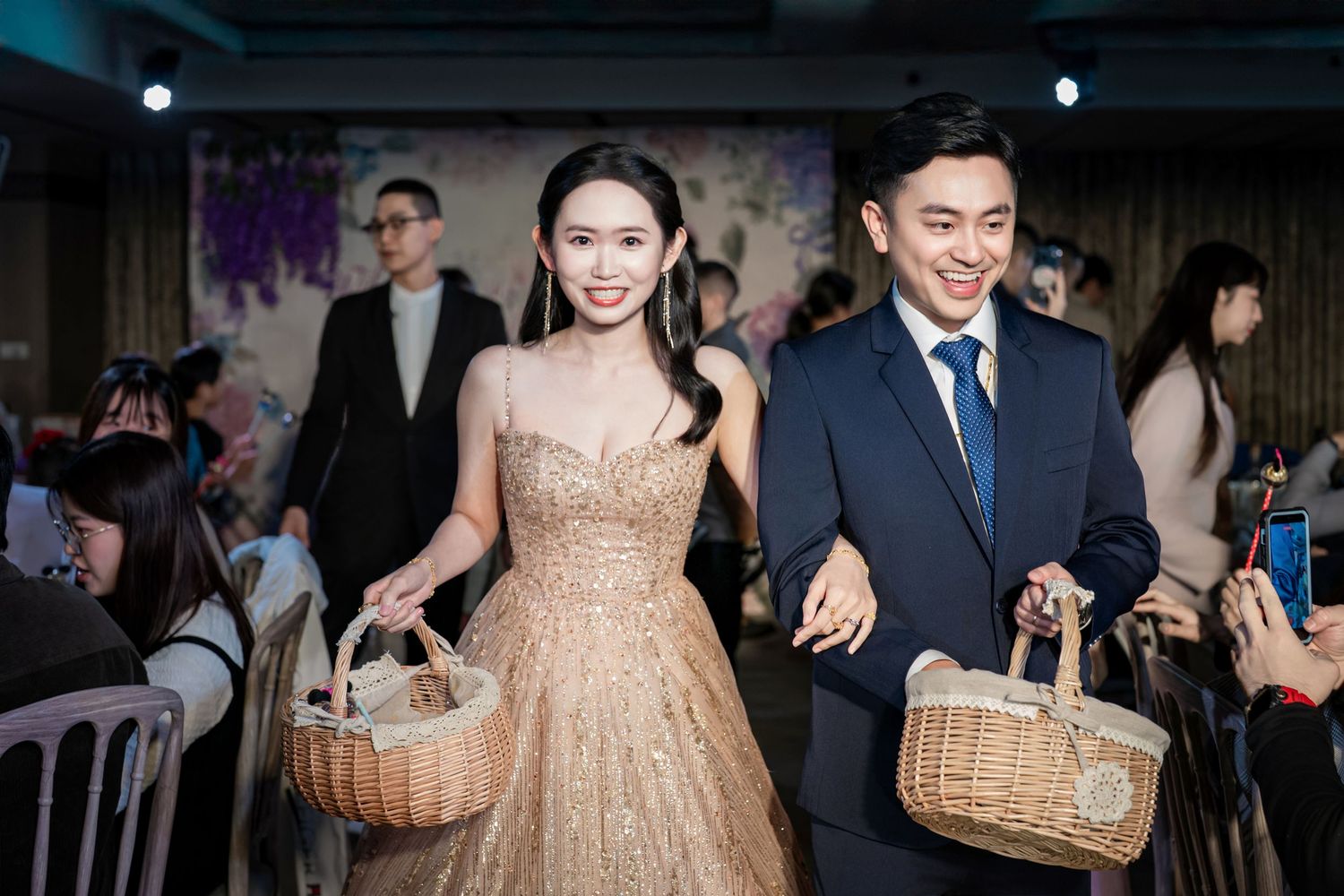 Elegantly dressed pair carrying woven baskets while walking together at an indoor celebration.