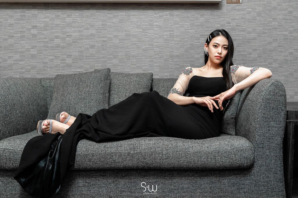 Elegant woman in black dress poses gracefully on a modern gray couch.