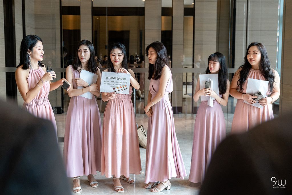 Bridesmaids in pink dresses gather together while holding papers during wedding preparations.