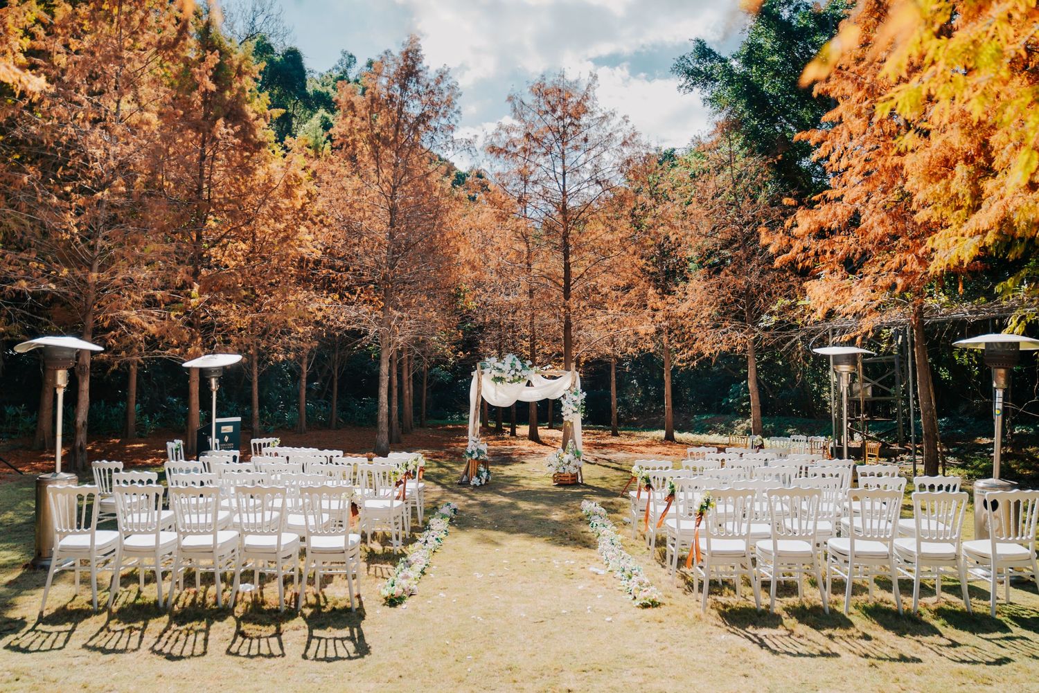 Outdoor wedding ceremony site with rows of white chairs set up between fall trees with golden leaves.