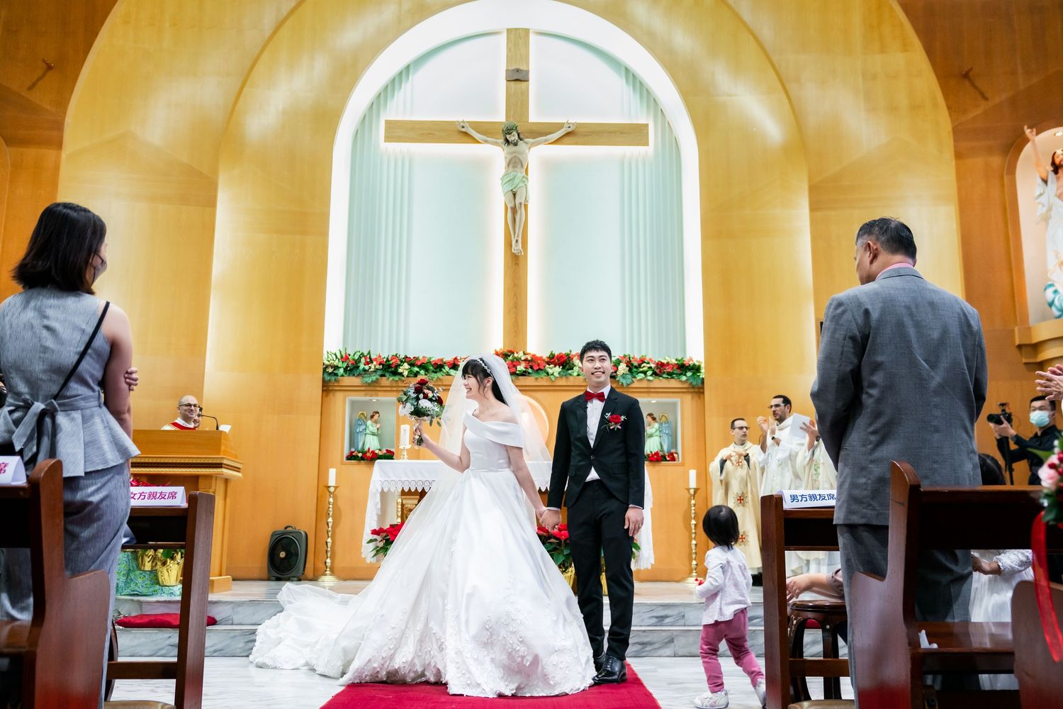 Newlyweds exit down a red-carpeted aisle in a yellow-walled church with a large crucifix.