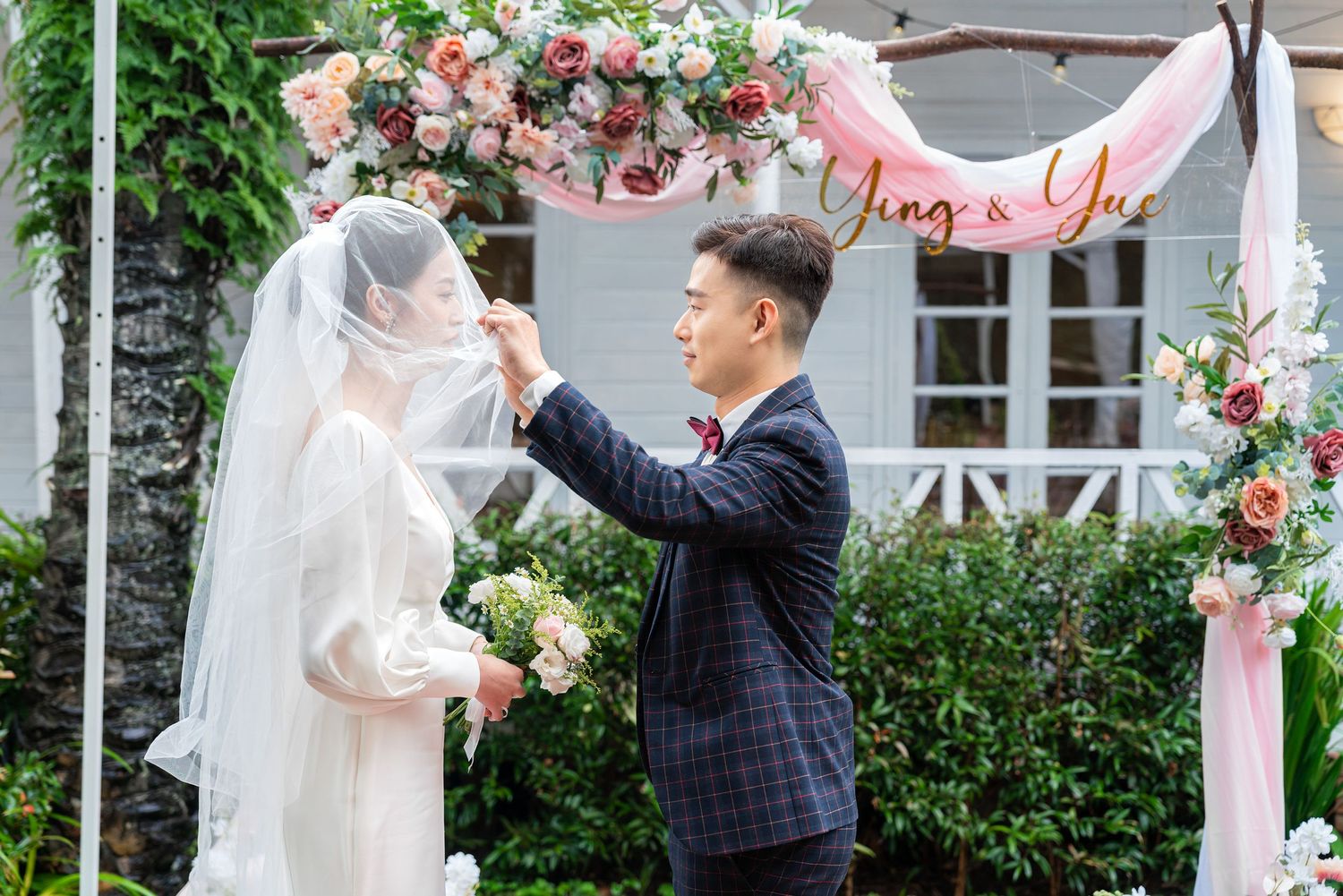 A wedding ceremony takes place under a floral arch with pink and white roses and natural greenery.