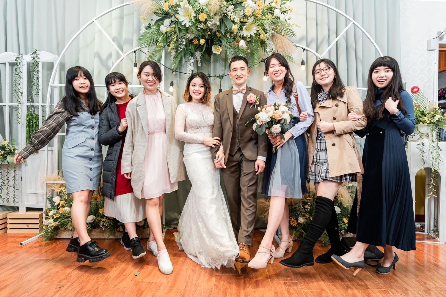 A group of friends pose together at an elegant indoor wedding celebration with floral decorations and arched backdrop.