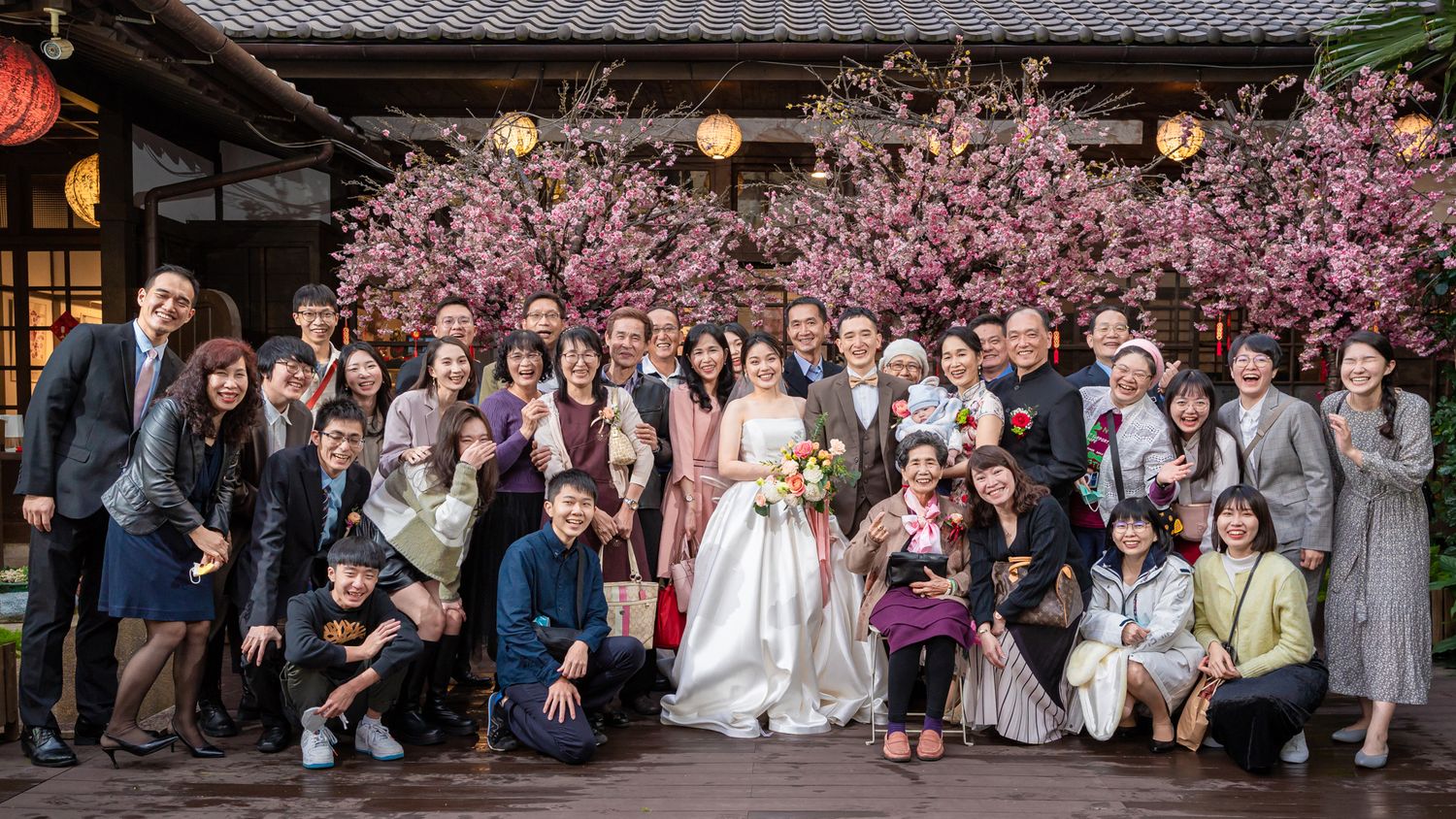 Wedding party gathers for group photo in front of cherry blossoms and traditional Japanese building.