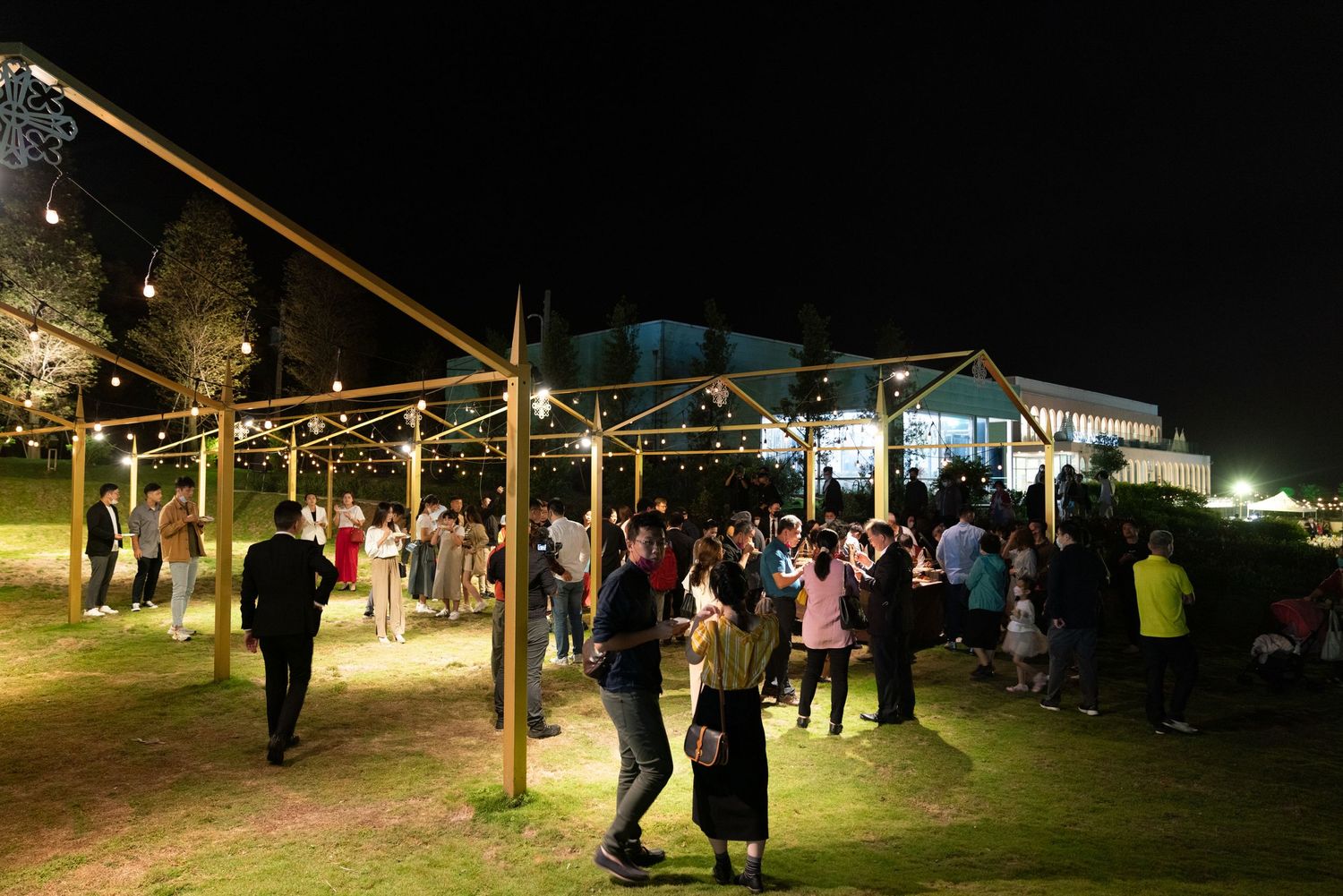 People gathering under string lights at an outdoor evening event with a large building in the background.