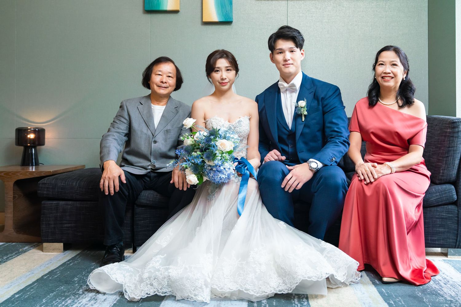 A wedding group sits on a modern couch with the bride in white holding blue flowers and guests in gray and coral attire.