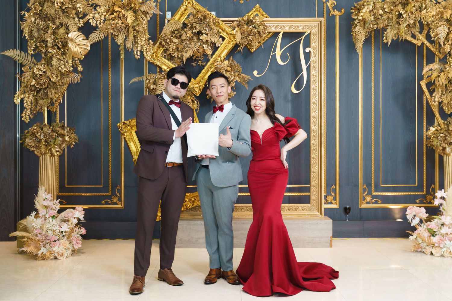 Three people posing together in formal attire against an ornate gold and blue backdrop with floral decorations.