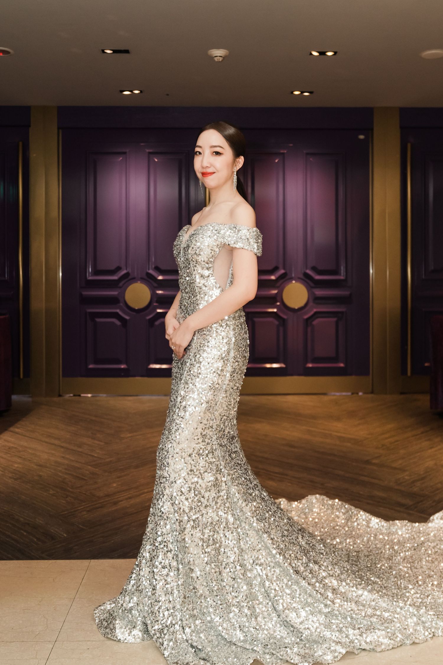 A person wearing a glamorous off-shoulder silver sequined gown with long train stands elegantly against purple doors.
