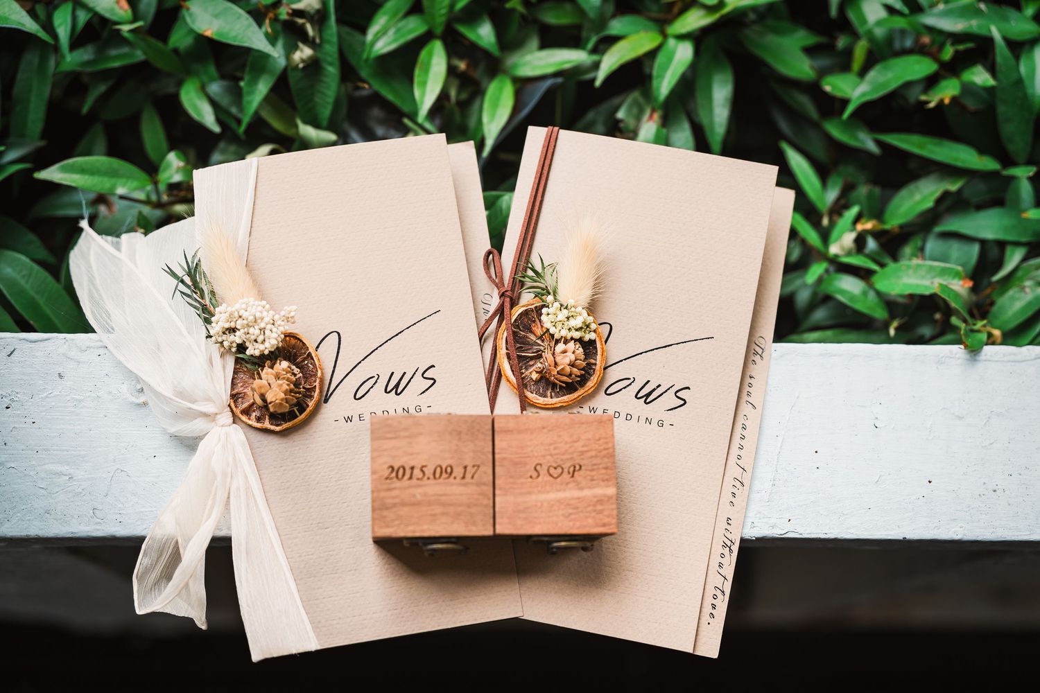 Two leather-bound vow books with gold detail and ribbon accents sit against a green leafy background.