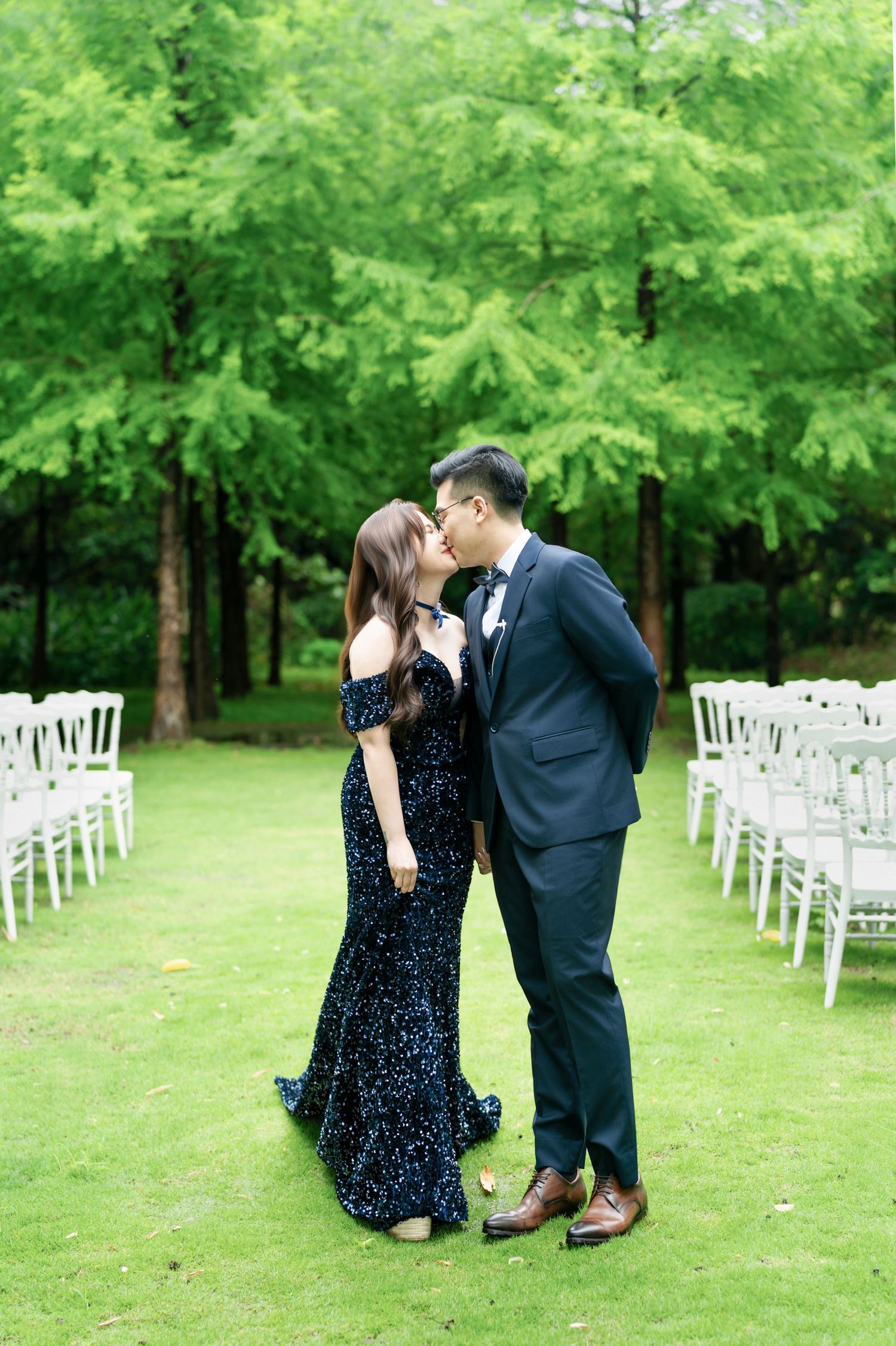 A romantic outdoor wedding setup with rows of white chairs on green grass surrounded by lush trees.