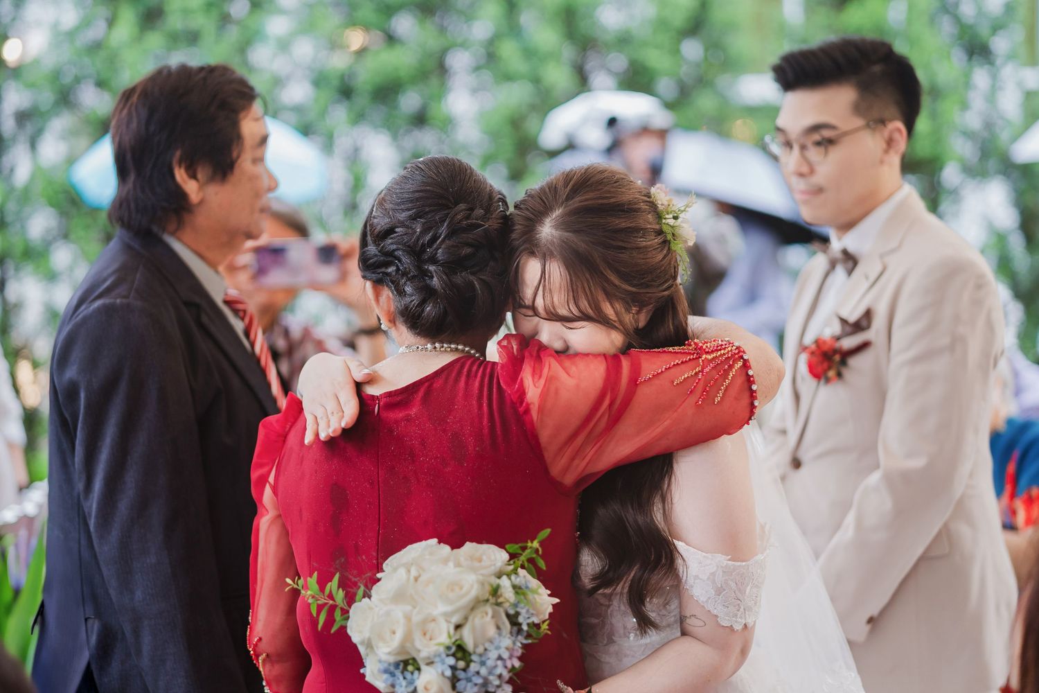 Emotional group embrace during an outdoor wedding ceremony with white floral bouquet.