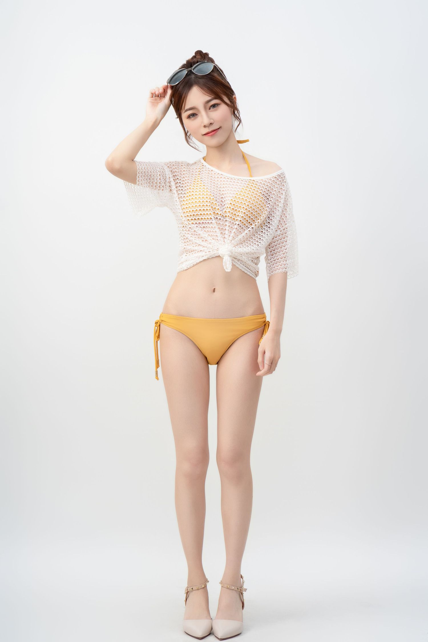 A woman in a white mesh top and yellow bikini bottom poses against a white backdrop in a stylish beachwear look.