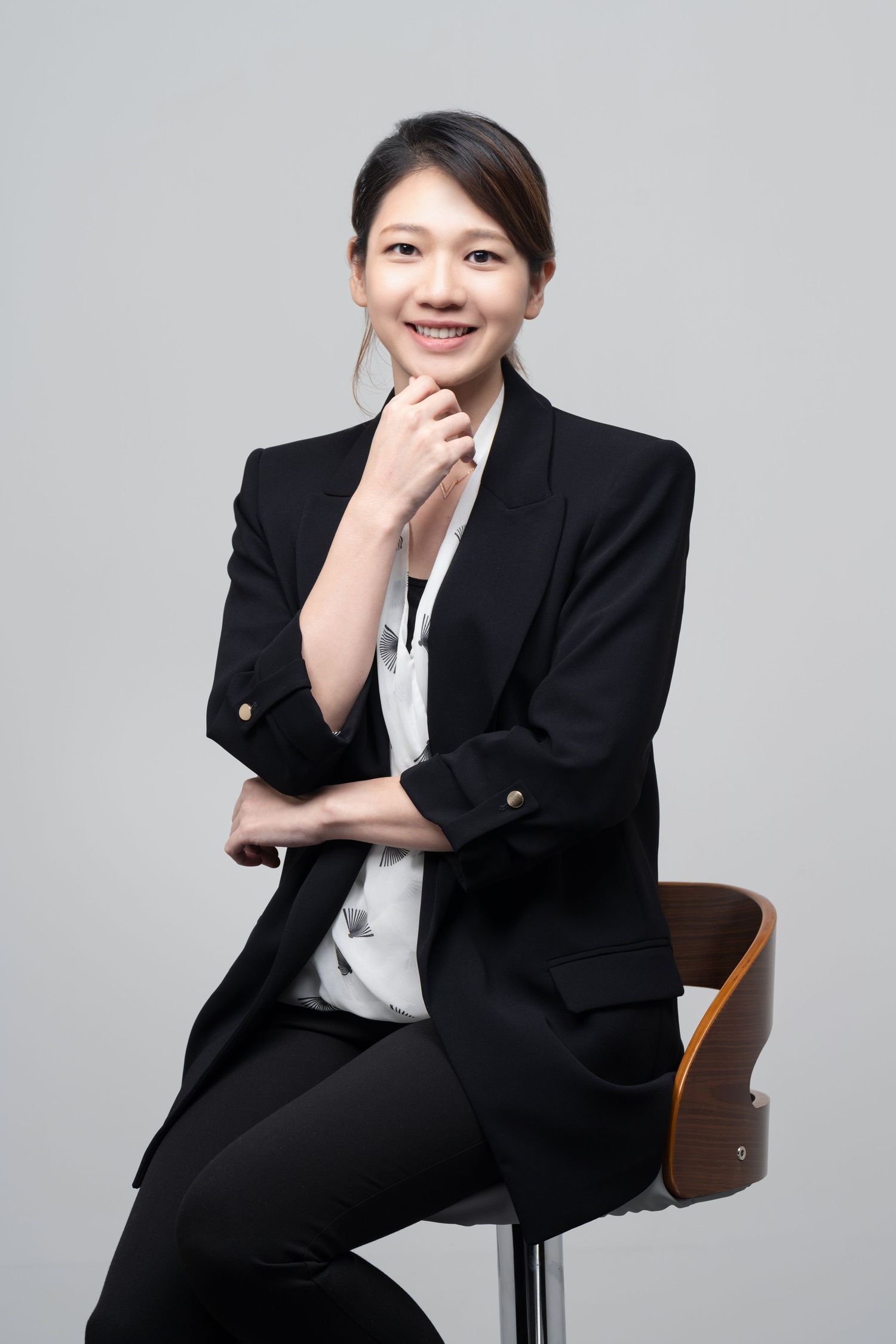 Professional studio portrait showing a person wearing a black blazer and white shirt seated casually on a wooden chair.