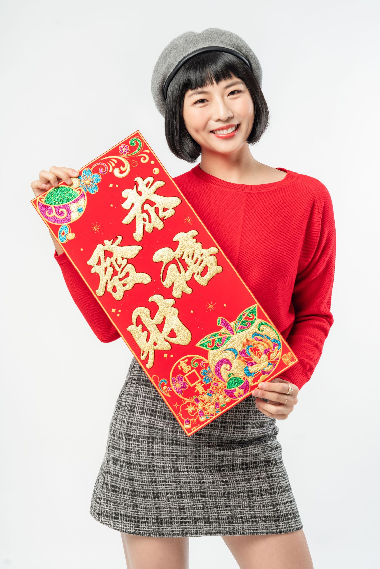 A person wearing a grey beret and red sweater holds up a traditional Chinese New Year red banner with gold text.