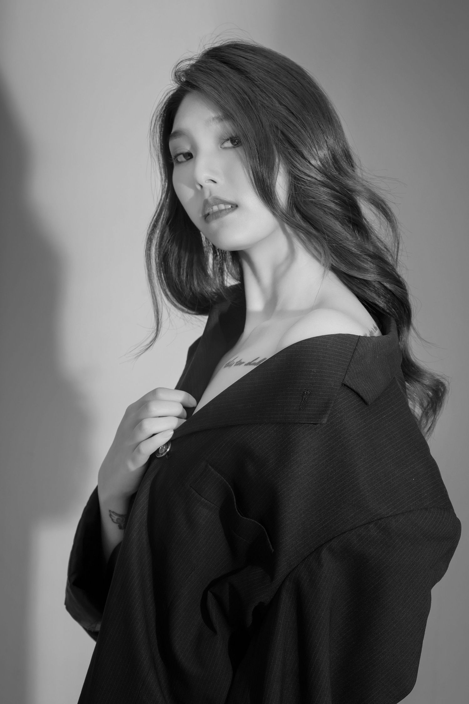Artistic black and white portrait of person wearing loose black blazer against plain backdrop with dramatic shadows.