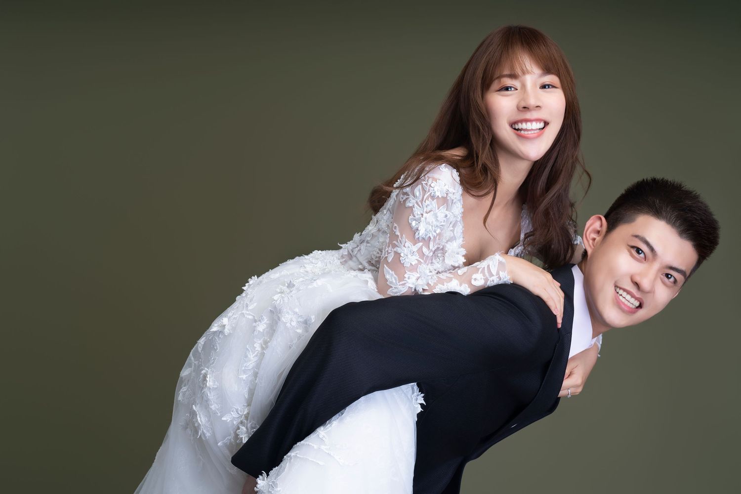 A couple poses playfully in wedding attire with dark green background as one person carries the other.