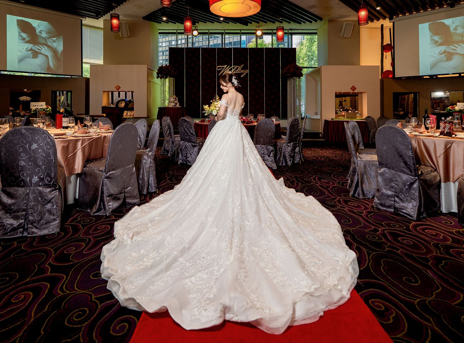 A wedding dress train flows elegantly across a red carpet in a dimly lit reception venue.