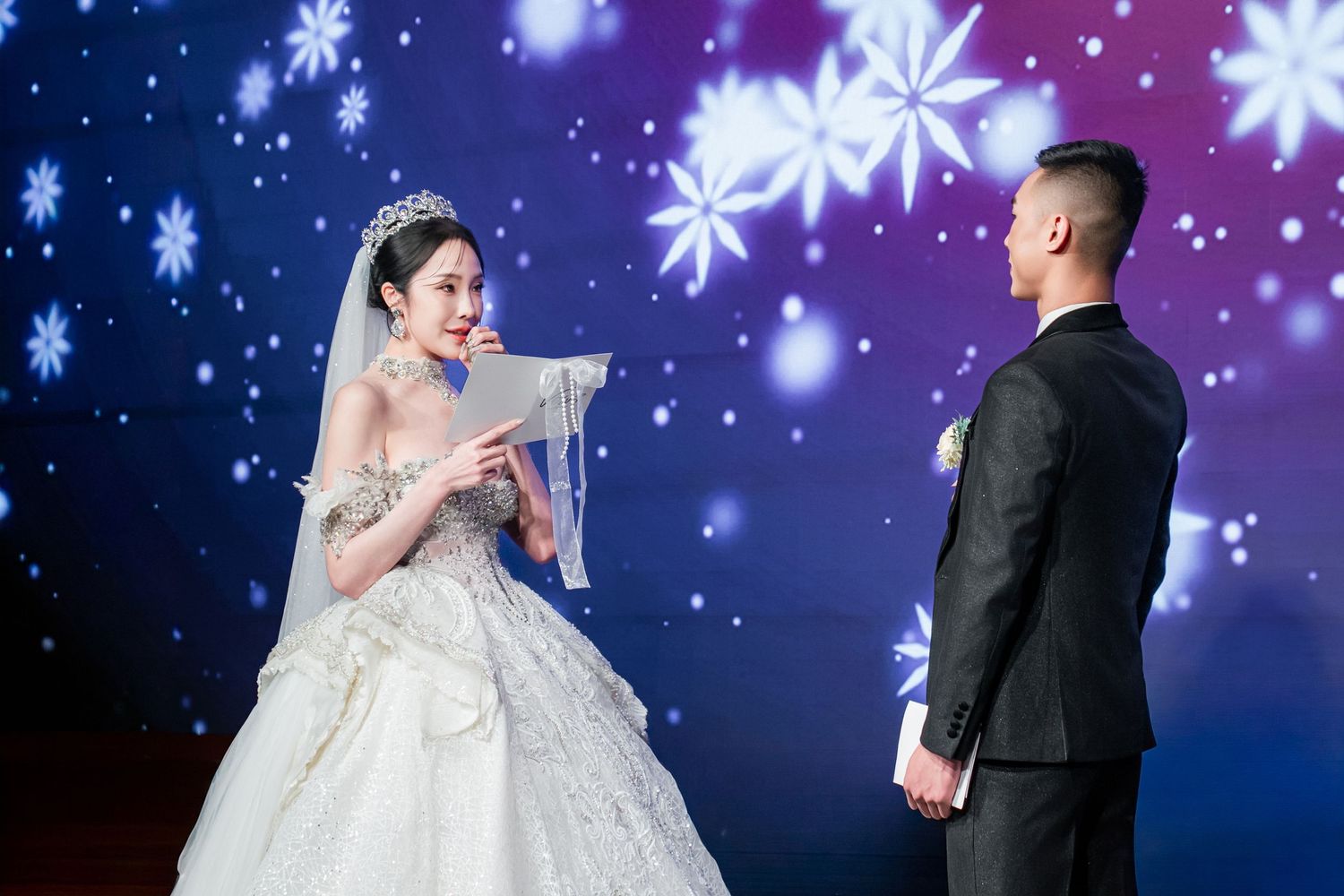 A couple in wedding attire poses against a starry blue and purple backdrop with snowflake decorations.