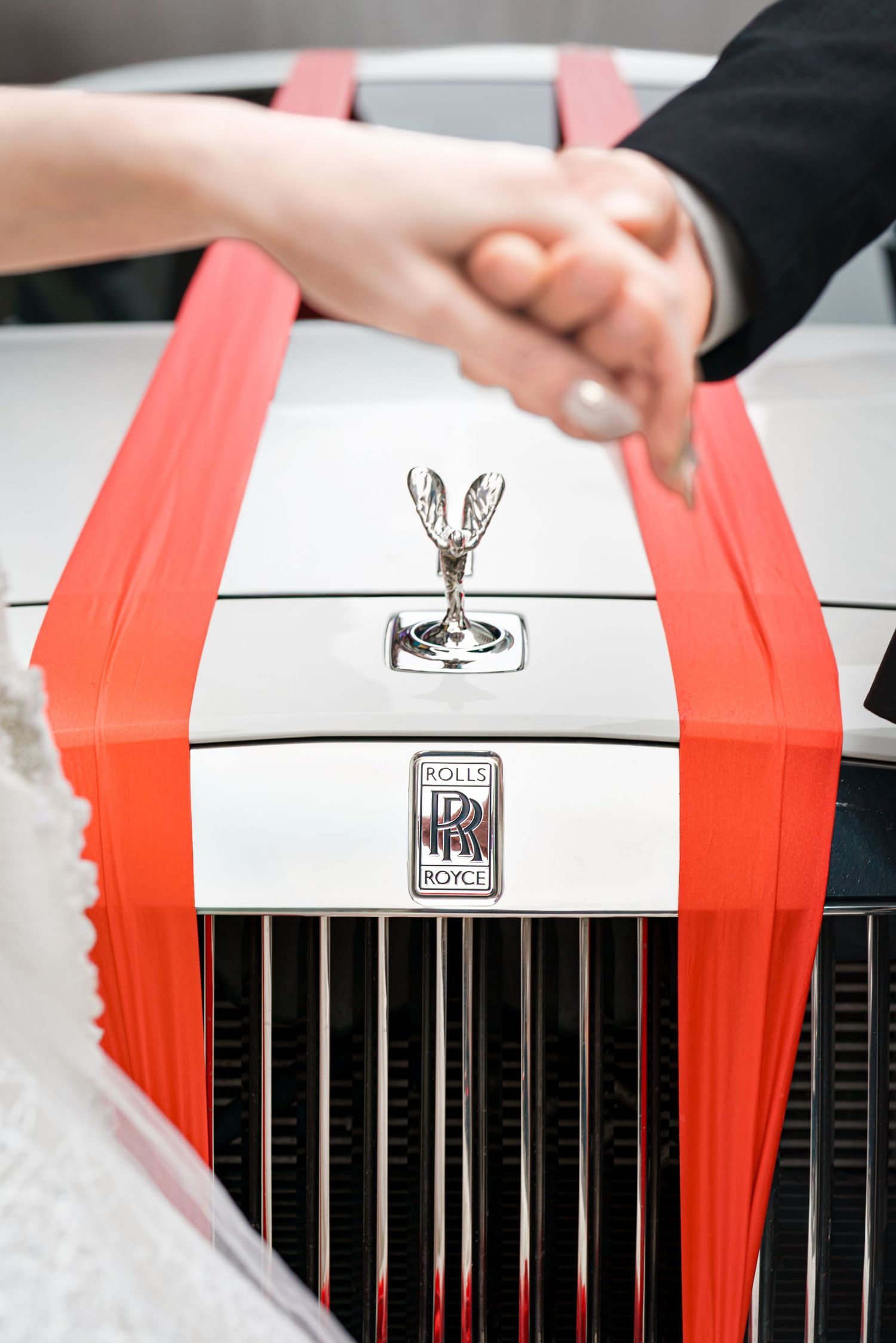 A white Rolls-Royce hood with iconic Spirit of Ecstasy ornament and red racing stripes, with hands clasped above.