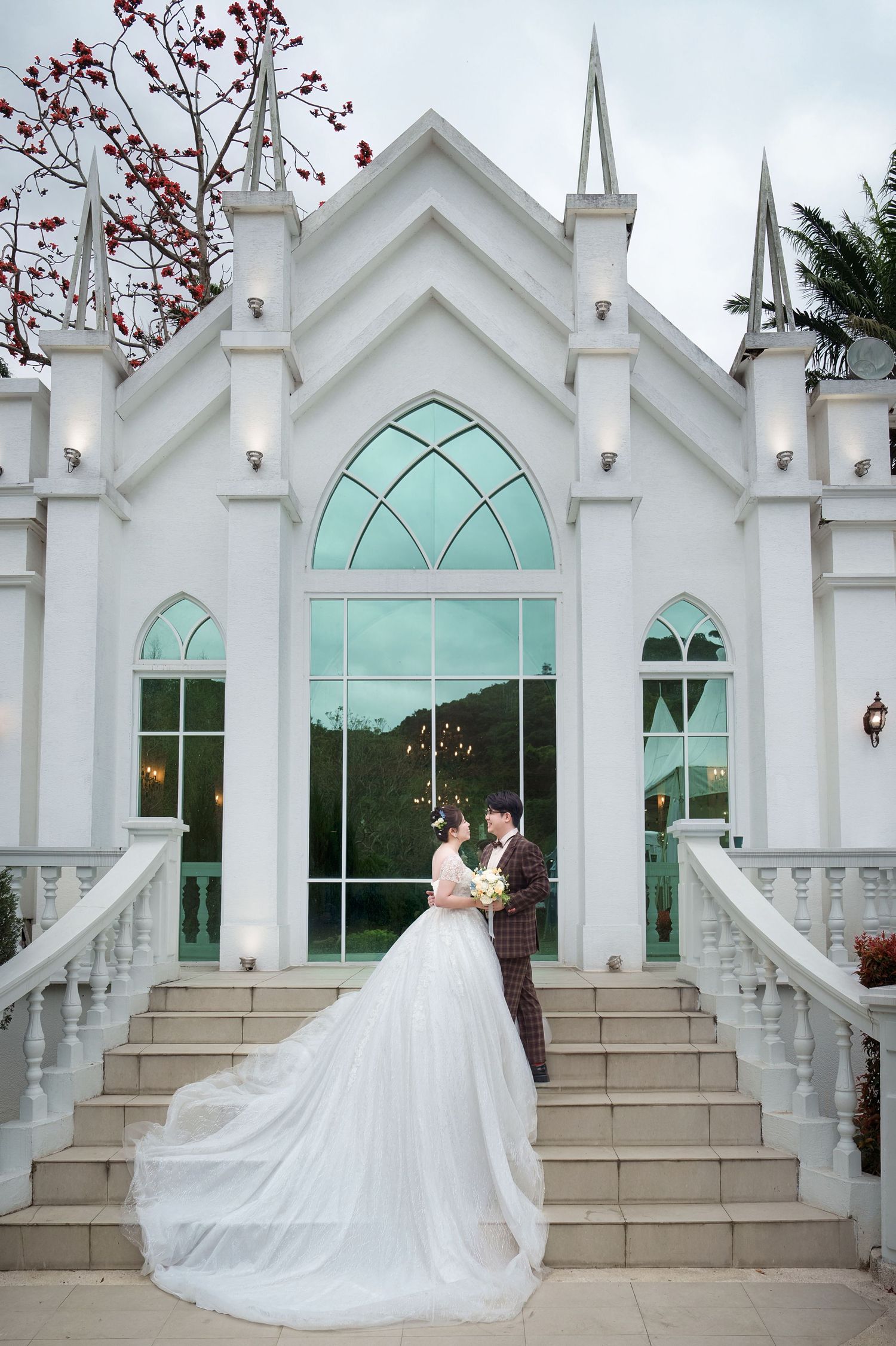 A bride in a flowing white wedding dress stands on stone steps in front of a white chapel with mint green arched windows.