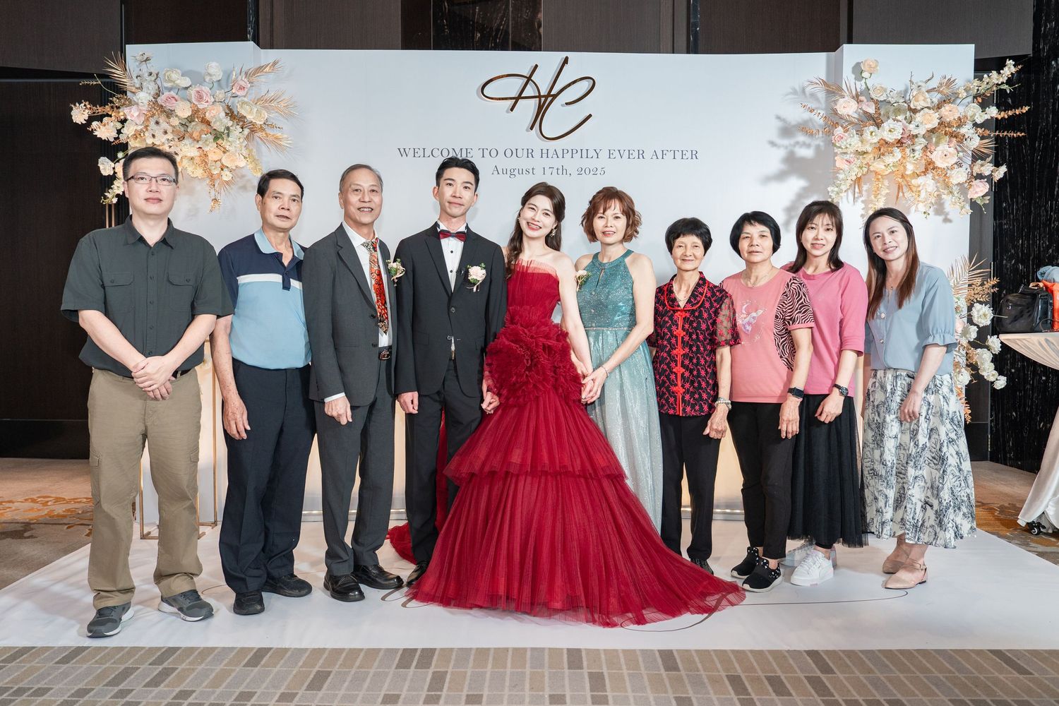 Wedding party poses together at celebration against backdrop with red formal gown as centerpiece.
