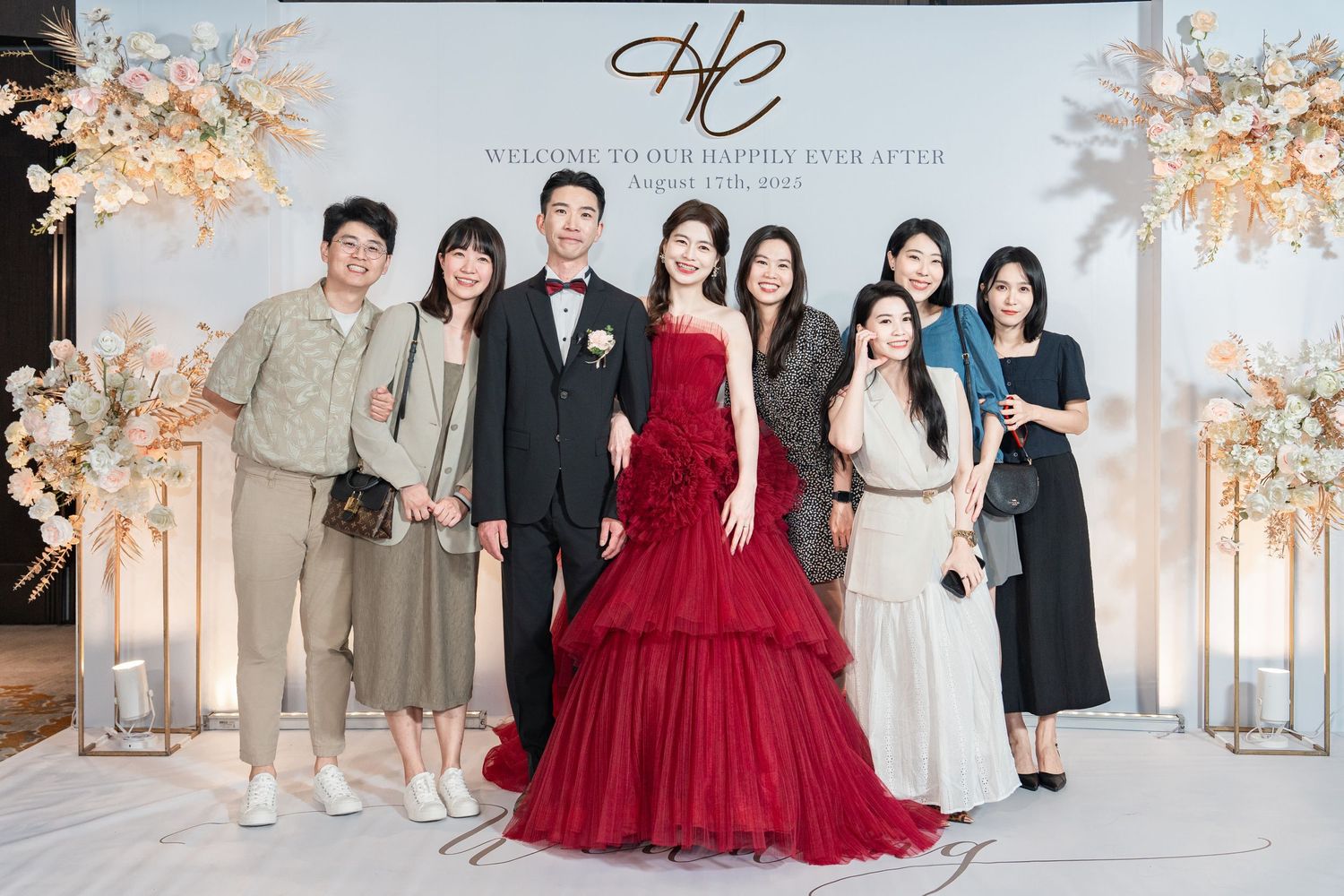 Elegant wedding celebration with guests posing by floral arch decor, featuring bride in stunning red ball gown.