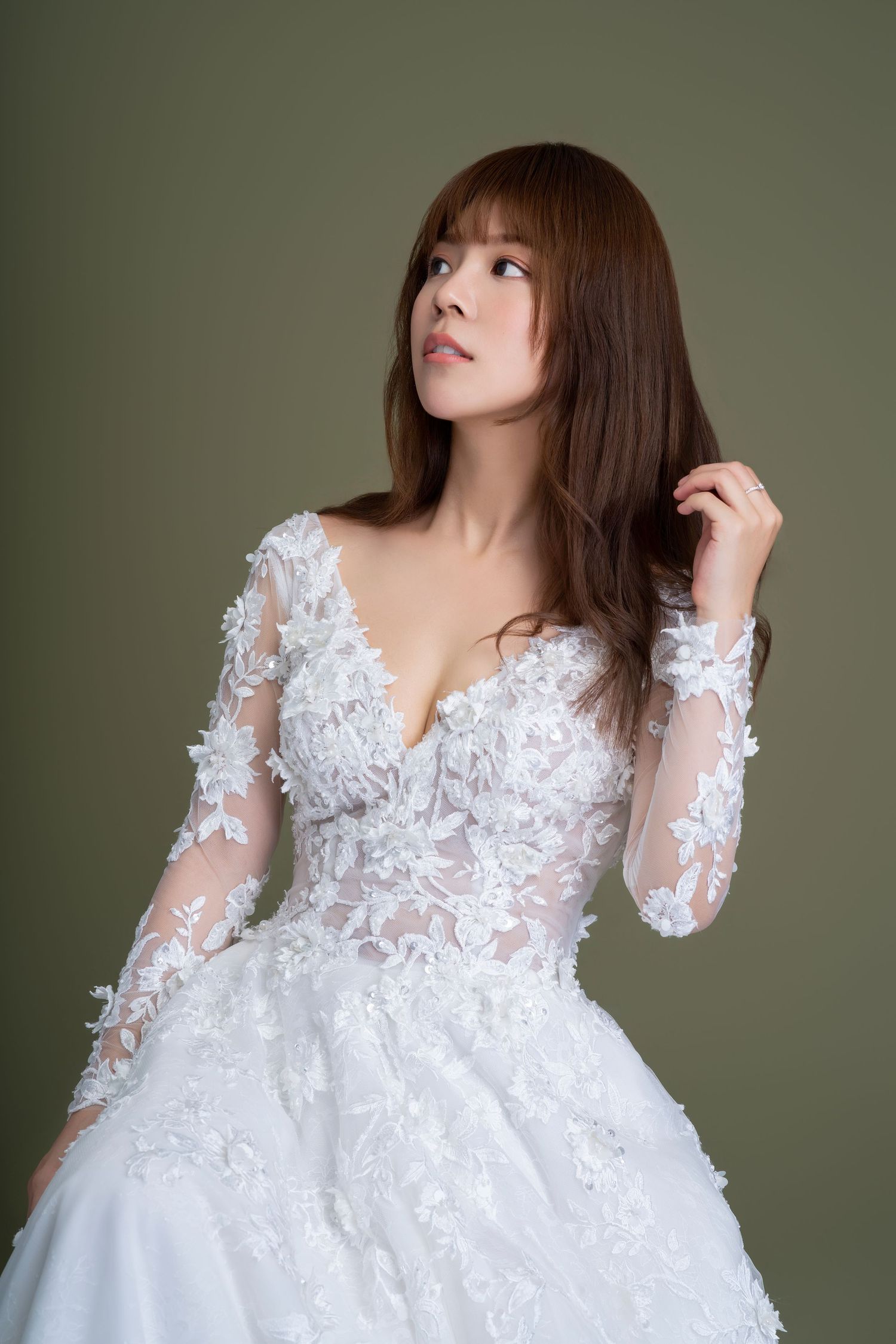 A white lace wedding dress with sheer long sleeves and floral appliques photographed against an olive green backdrop.