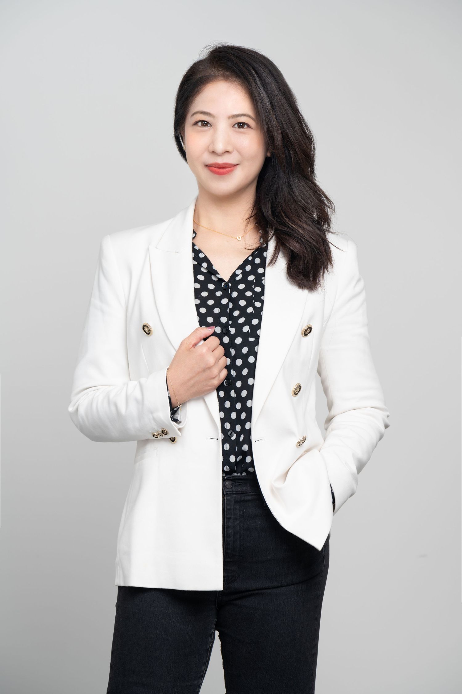 Professional portrait of someone wearing a crisp white blazer with polka dot blouse against a gray background.