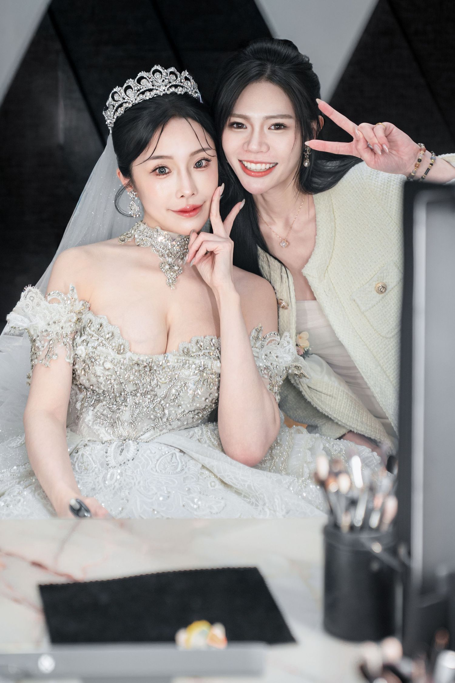 A bride in a sparkling white wedding gown and tiara poses playfully with another person at a celebration.