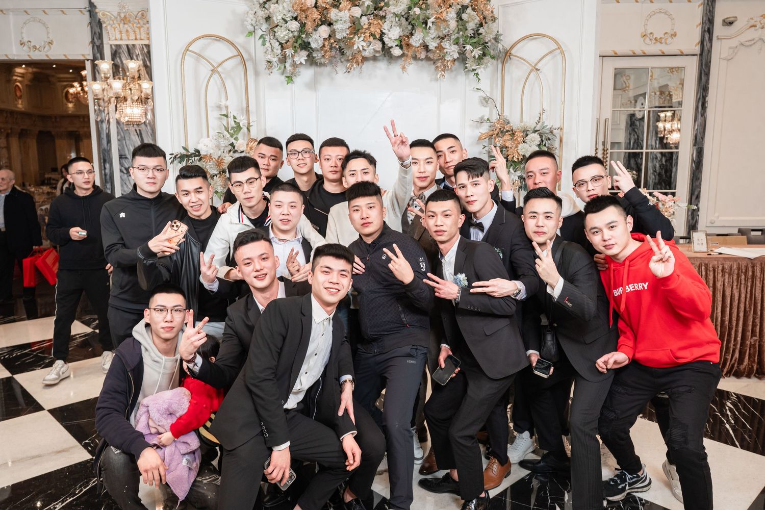 A large group in formal black suits pose together at an elegant venue with decorative floral arrangements.