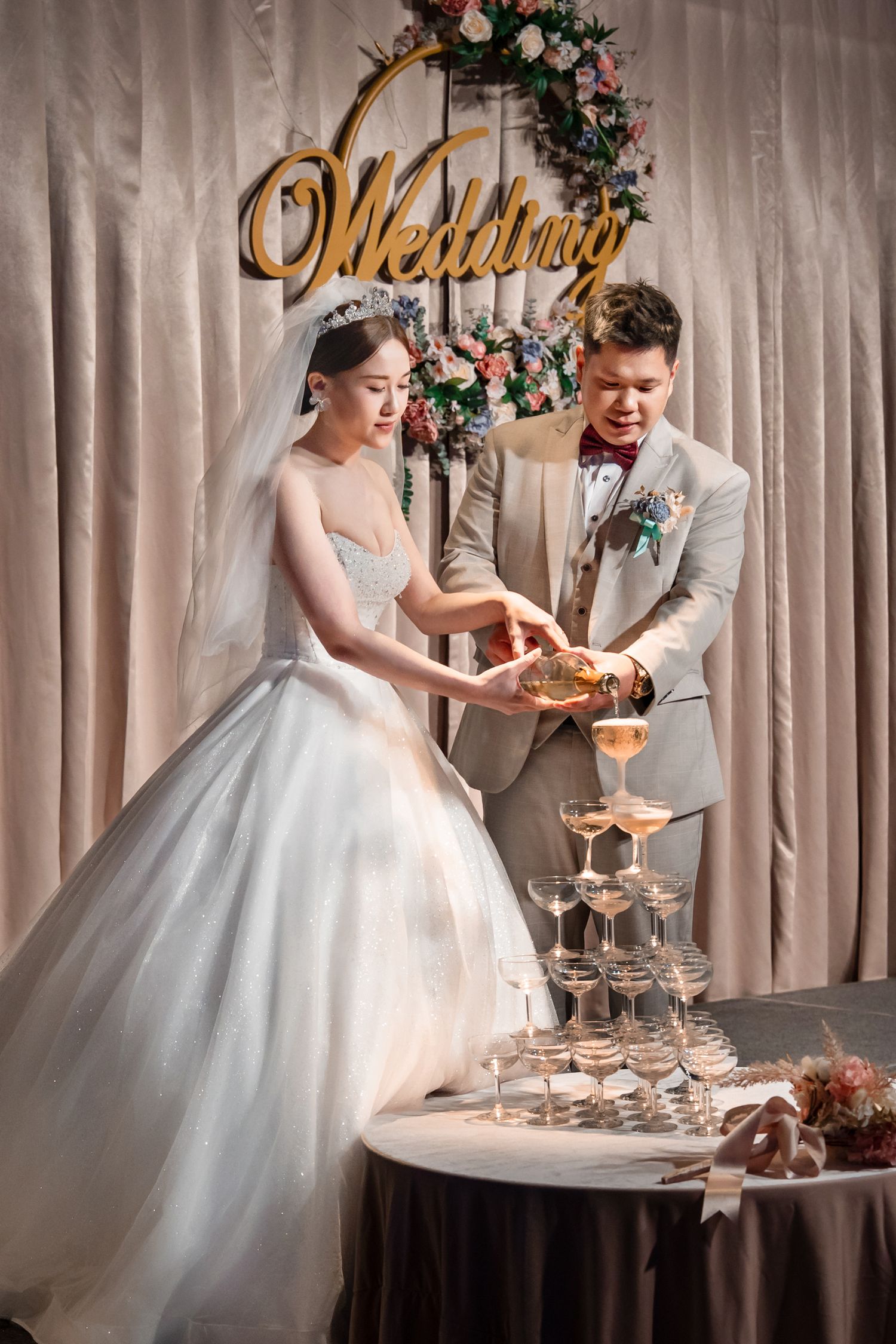 A couple in wedding attire arranges a tiered display of desserts at their reception table.