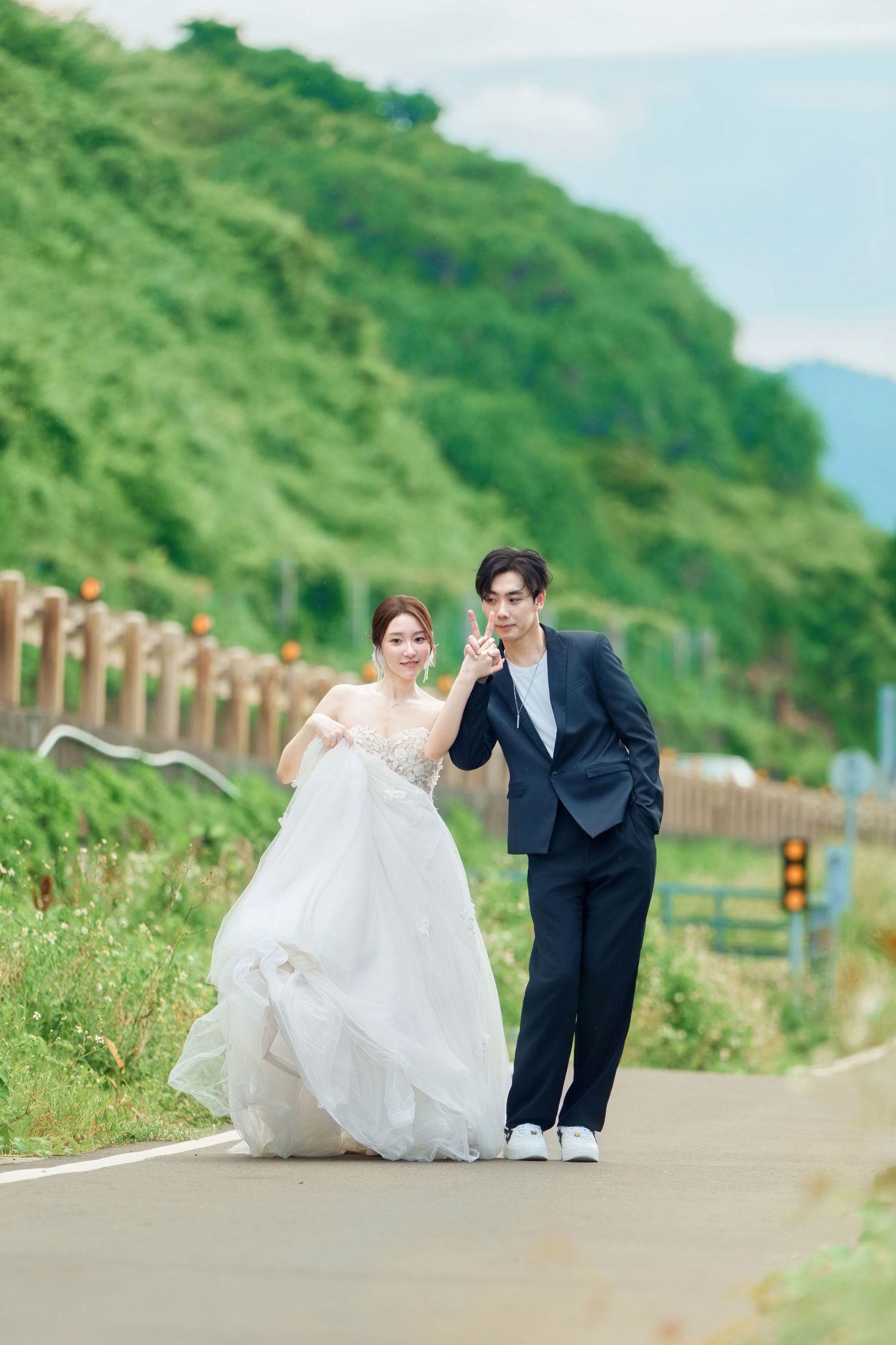 A couple in wedding attire walks along a scenic path with green hills and mountains in the background.