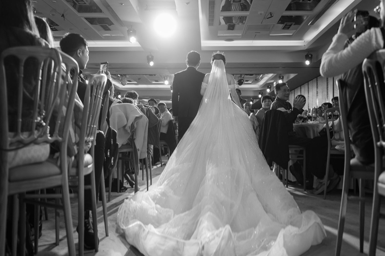 A bride in a flowing white wedding gown walks down a train aisle while passengers watch and smile.
