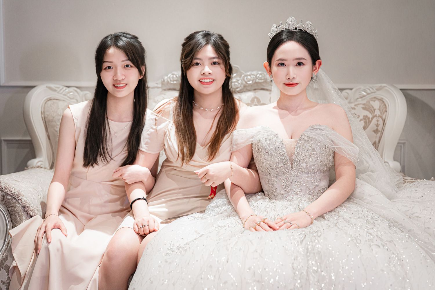 Three women in elegant dresses sit together on an ornate white sofa, with one wearing a beautiful wedding gown.