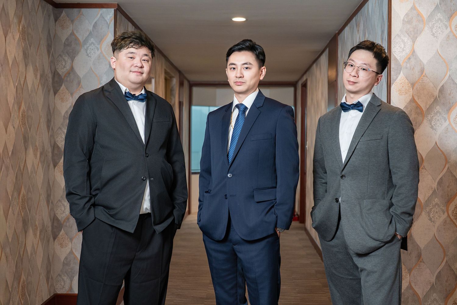 Three men in formal attire pose together in a hotel hallway wearing suits and bowties.