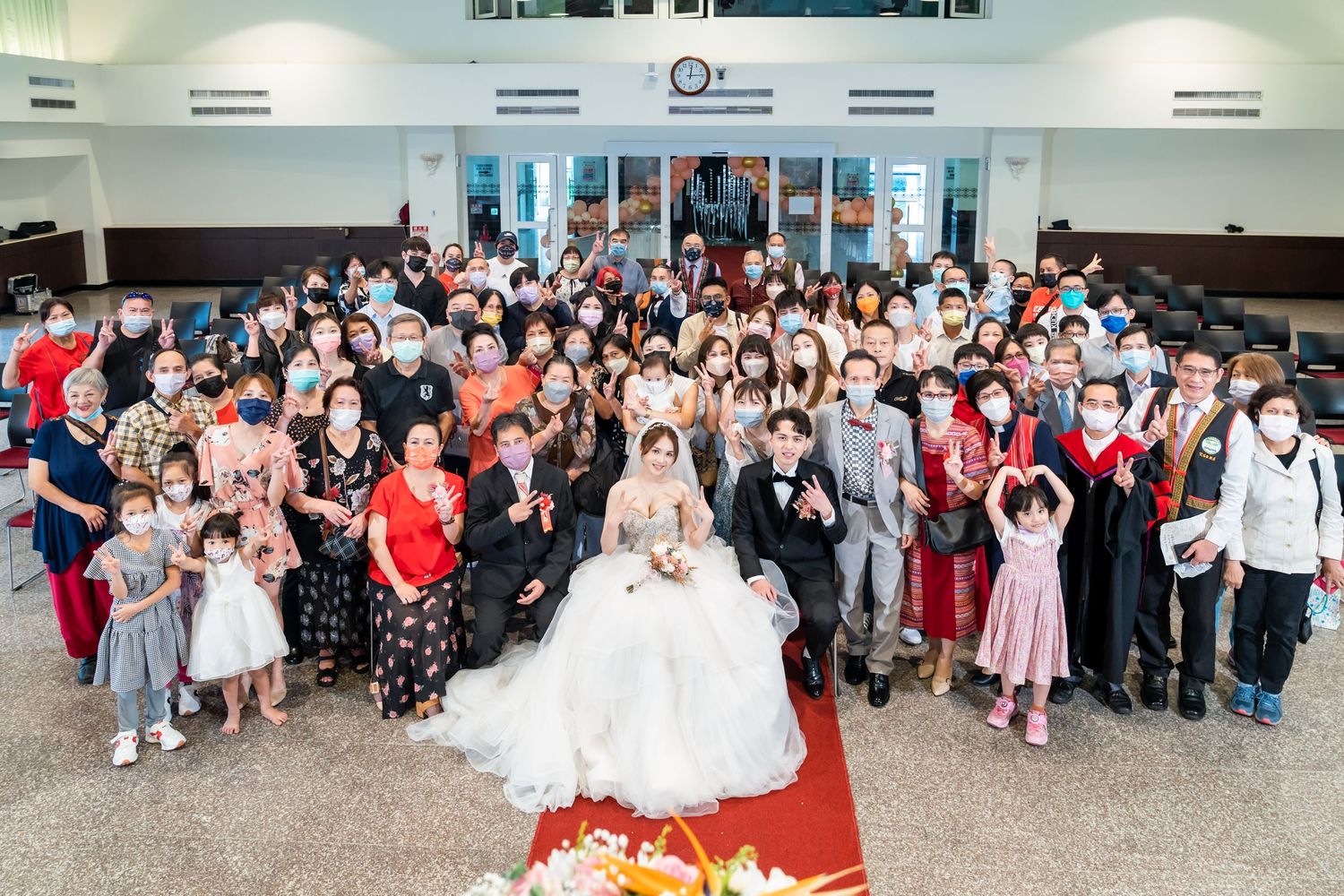 Large wedding group photo with guests gathered on red carpet in church hall with white walls.