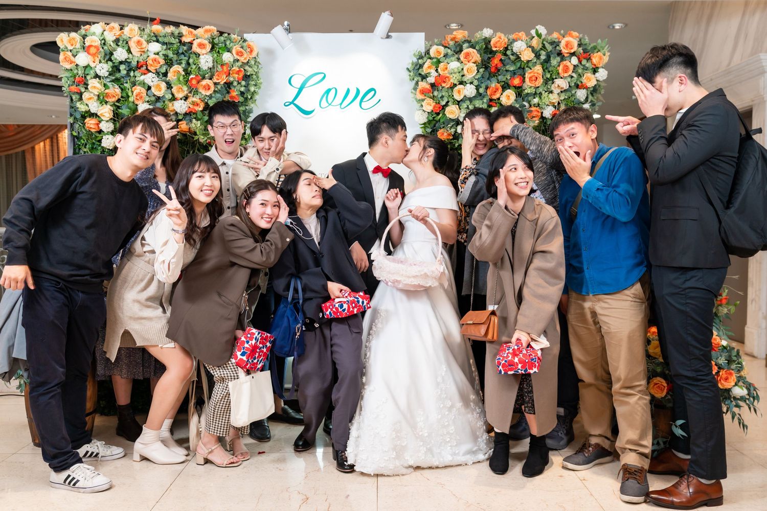 Wedding celebration group shot with happy guests and couple in front of floral backdrop.