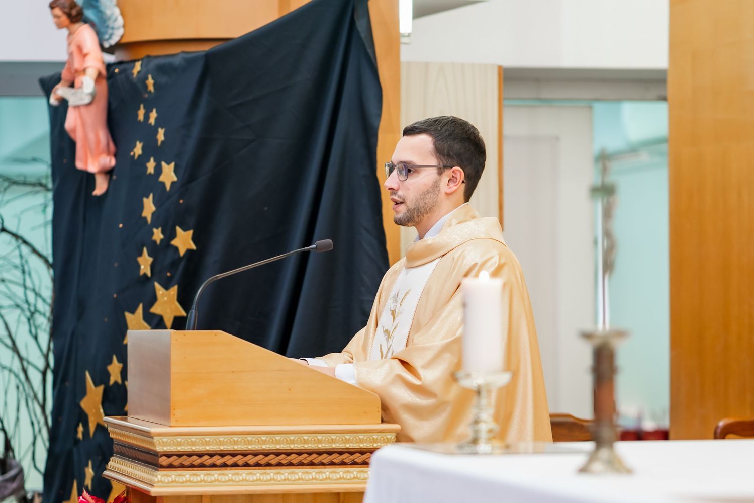 A speaker in cream colored robes stands at a wooden podium with a backdrop decorated with golden stars.