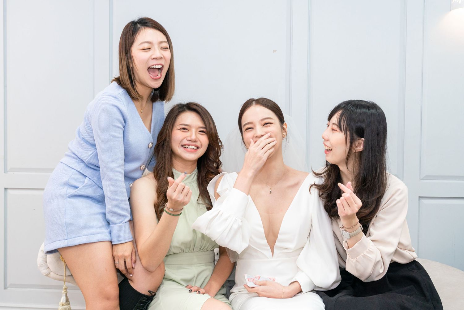 A group of women sitting together on a couch sharing laughs and covering their mouths while wearing casual attire.