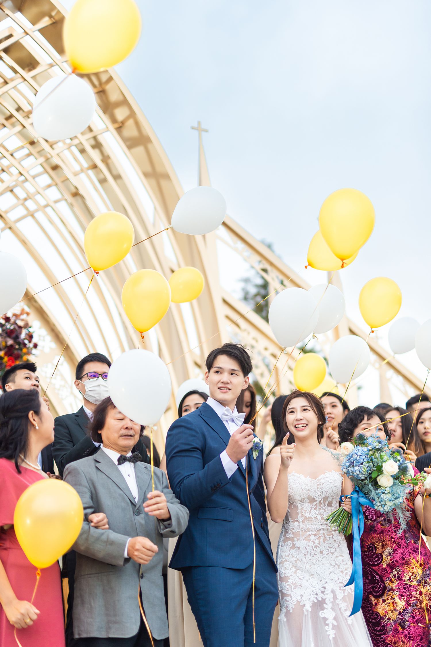 Wedding celebration outside a church with yellow and white balloons floating against an arched architectural backdrop.