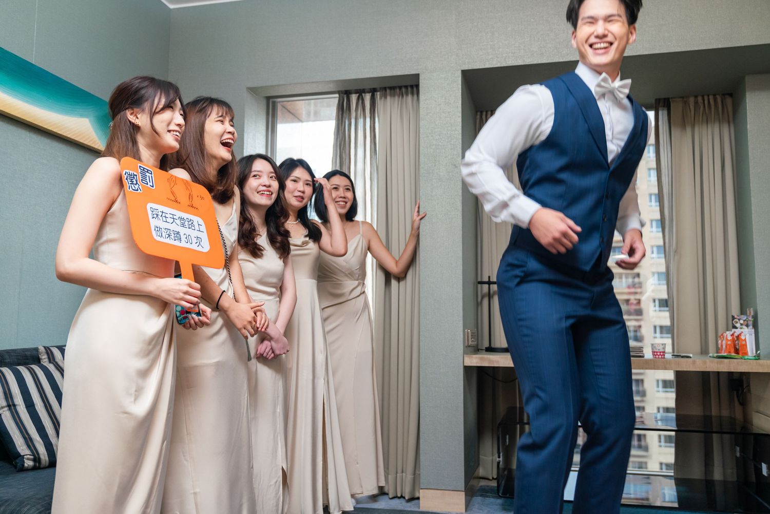 Wedding party members in neutral-colored dresses laugh together while a groomsman in blue vest poses separately.