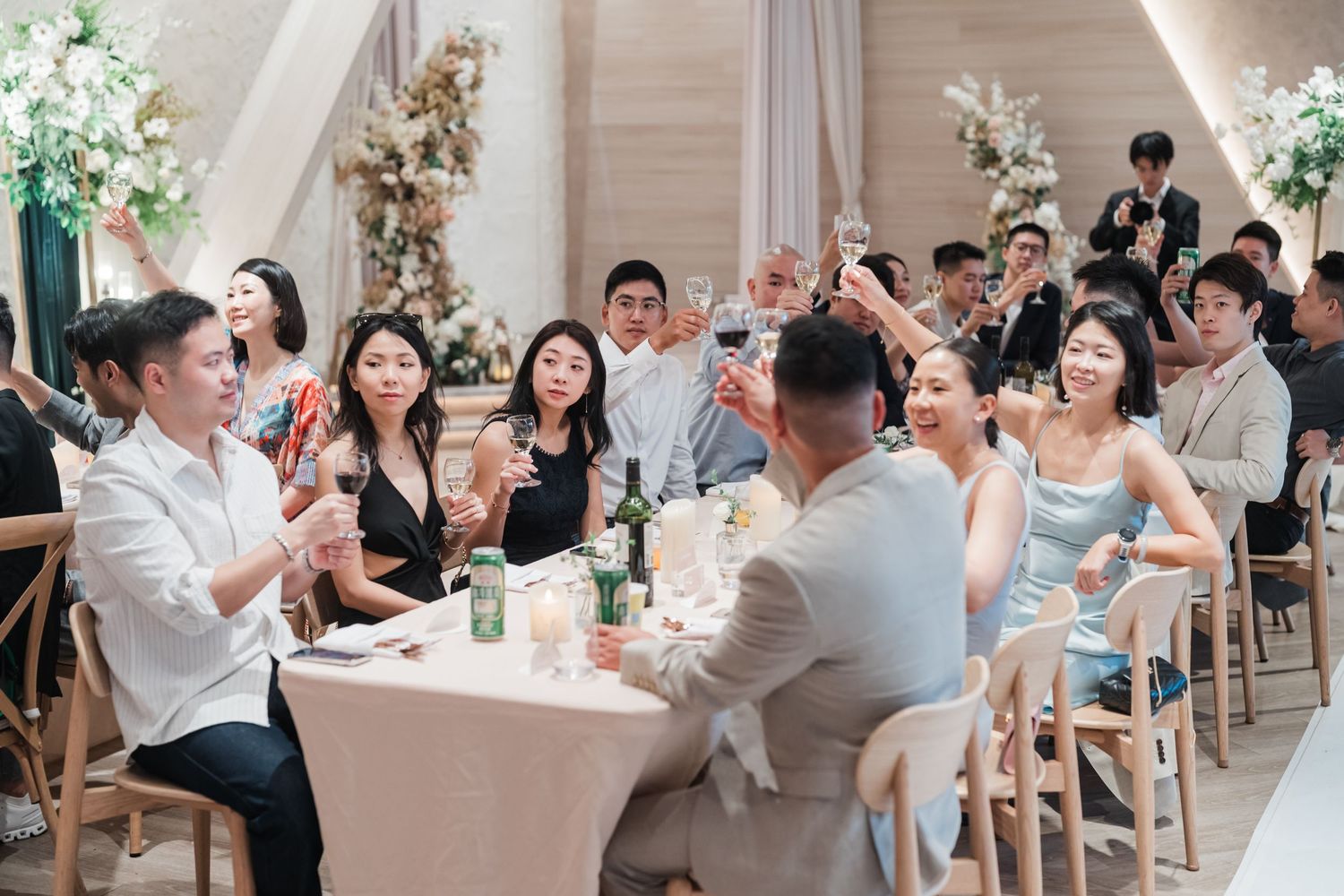 Wedding reception guests raise their glasses in celebration at an elegant indoor venue with white floral decorations.