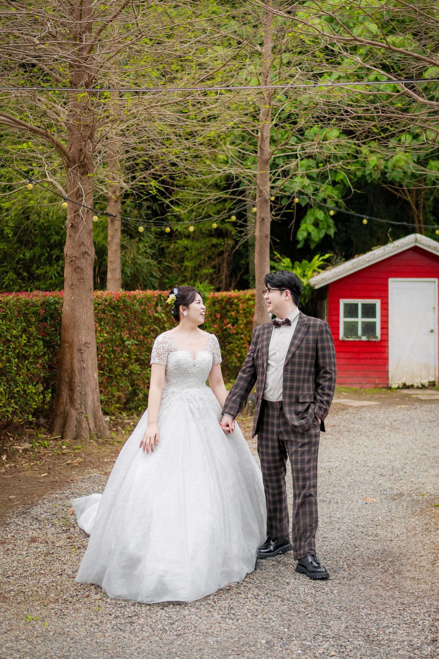 A couple in wedding attire stands together on a gravel path near a red barn and tall trees in an outdoor setting.