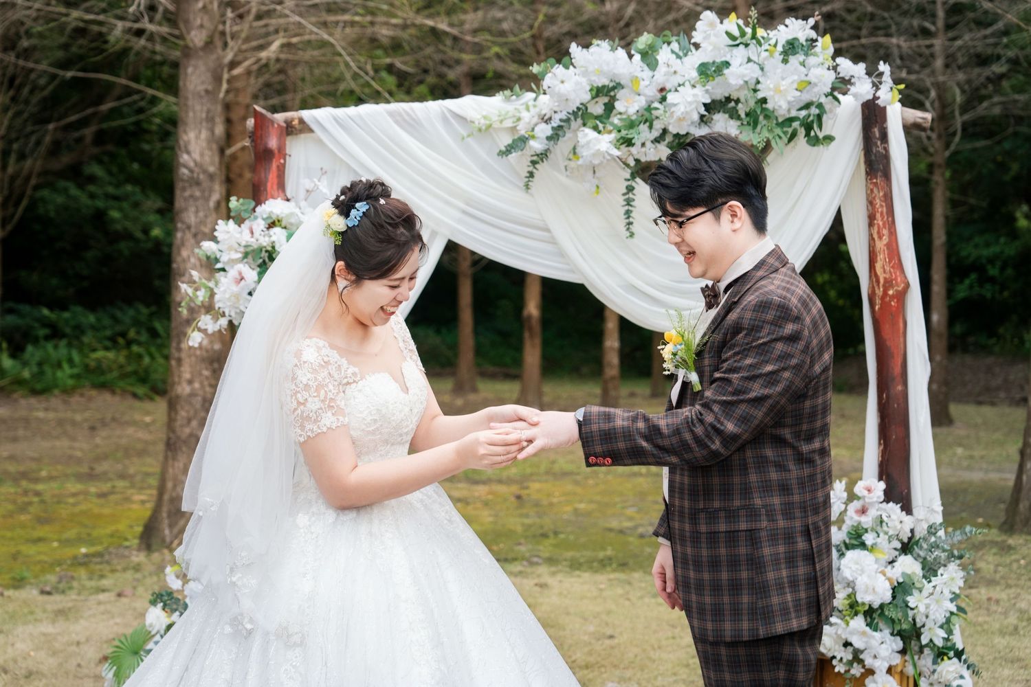 A romantic outdoor wedding ceremony takes place under a white fabric arch decorated with pale blue and white flowers.