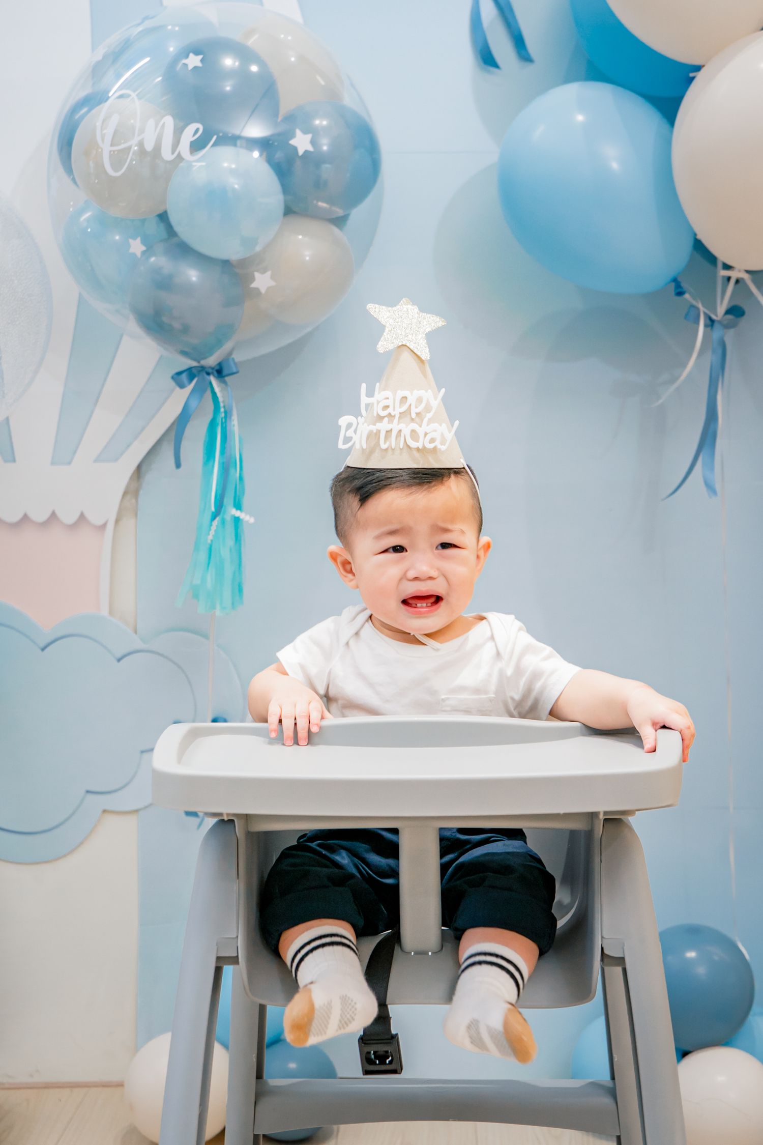 Baby sitting in grey high chair with blue and white balloons decorating background for first birthday celebration.
