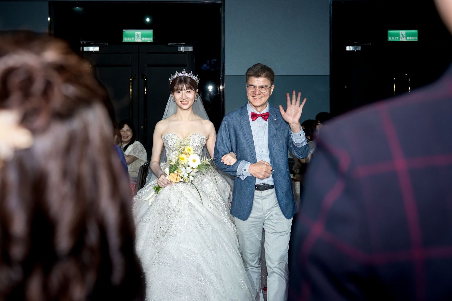 Bride in white gown and groom in blue suit waving at wedding reception entrance ceremony.