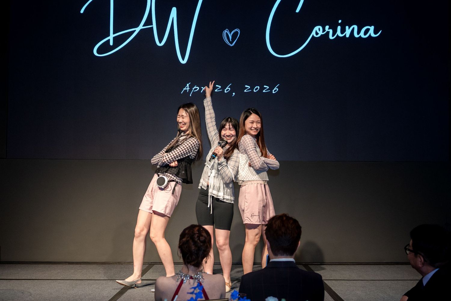 Three women posing on stage at a wedding reception with DW and Corina April 26, 2026 displayed behind them.