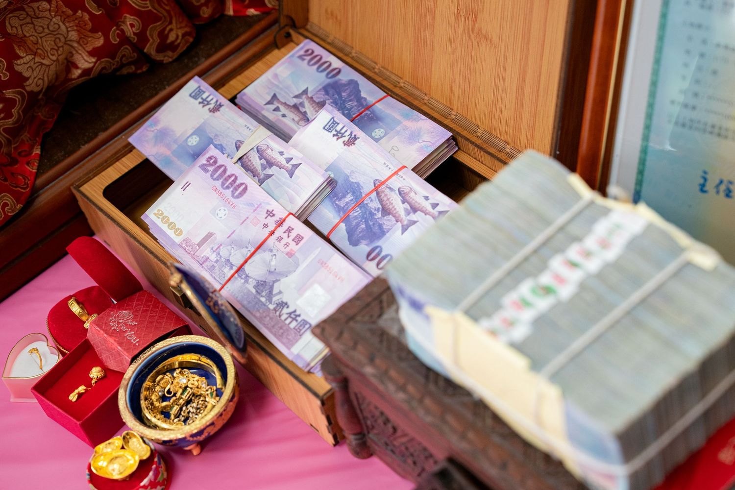 Stacks of New Taiwan Dollar banknotes bundled with red bands displayed in a wooden box with gold jewelry.