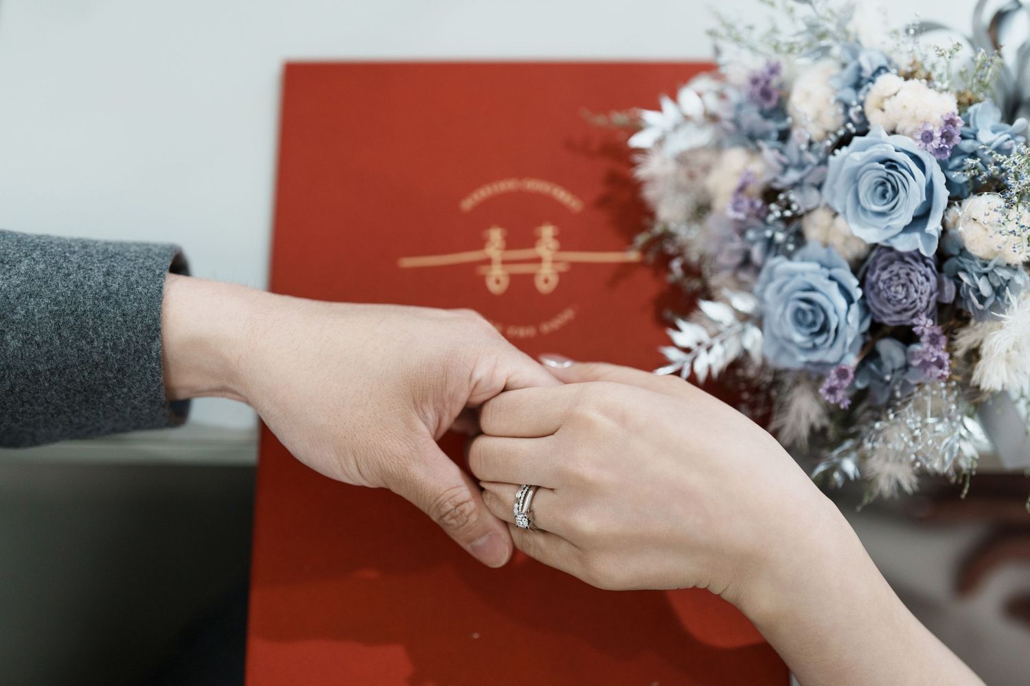 A sequence of hands exchanging a delicate blue and silver floral bouquet against a red background.