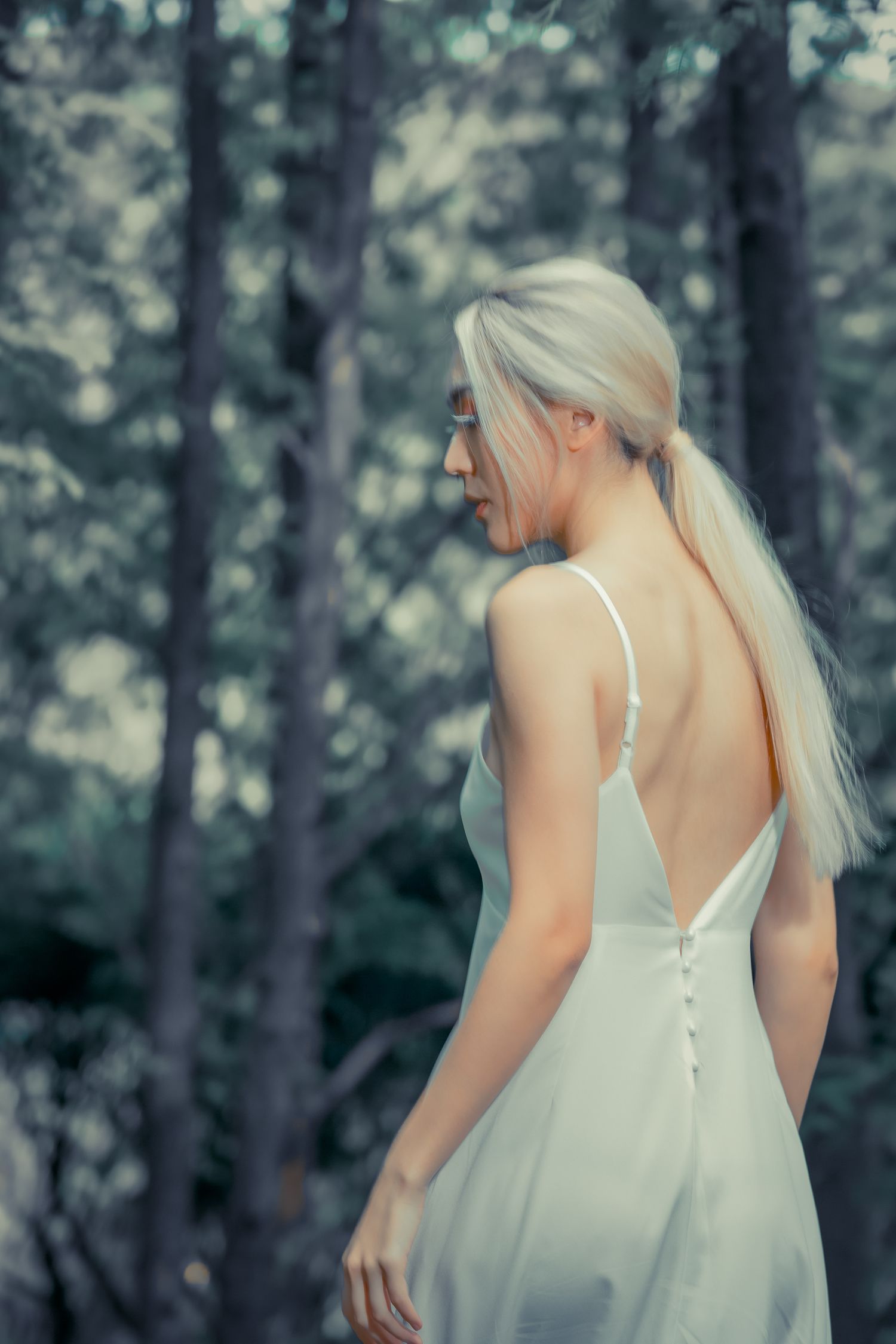 Person with long platinum blonde hair wearing a white dress with buttons stands in a misty forest setting.